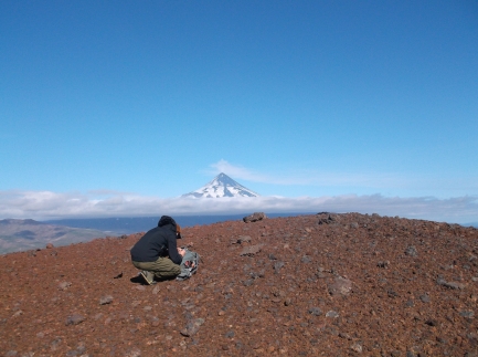 Man kneels to collect volcanic ash on a scenic overlook of a volcano