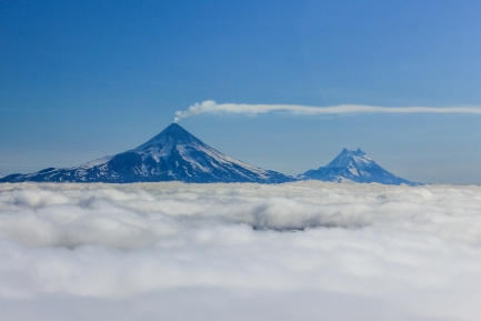 Two volcanoes, one with a stream of smoke coming out of the top, seen above a plume of clouds