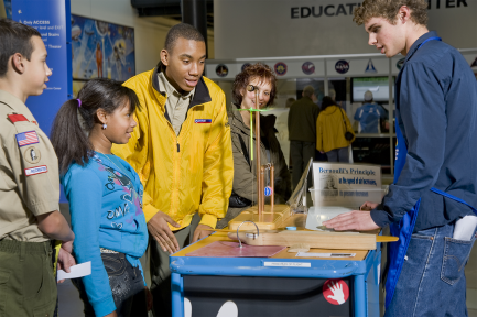 A young girl in a blue top stands next to a taller man in a yellow jacket in front of a rolling cart demonstration.
