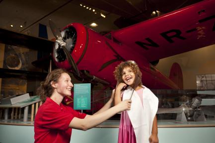 Museum Explainer Rose Agger helps a young girl dress up like a historic aviator