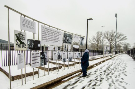 Person stands in snow looking up at art attached to a fence