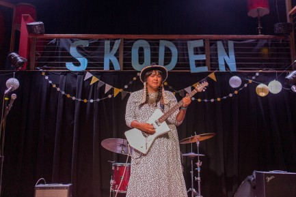 Woman performing on stage with guitar, SKODEN banner behind her