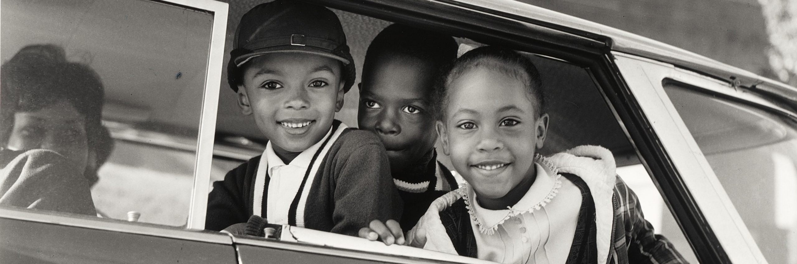 Three children smile from a car window on their way to school